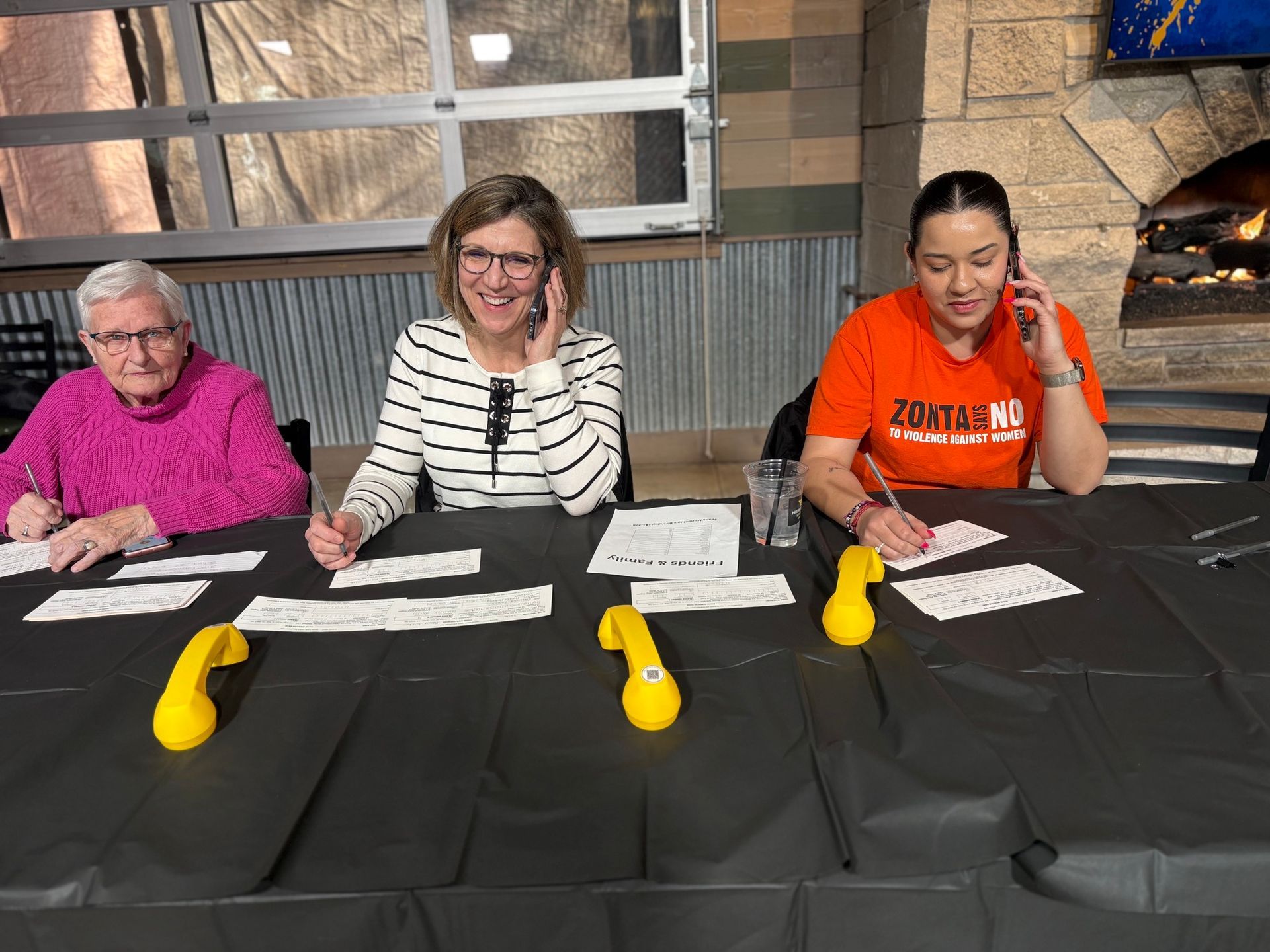 Three women on phones at a table. One in pink, one in stripes, and one in orange. All smiling and writing.
