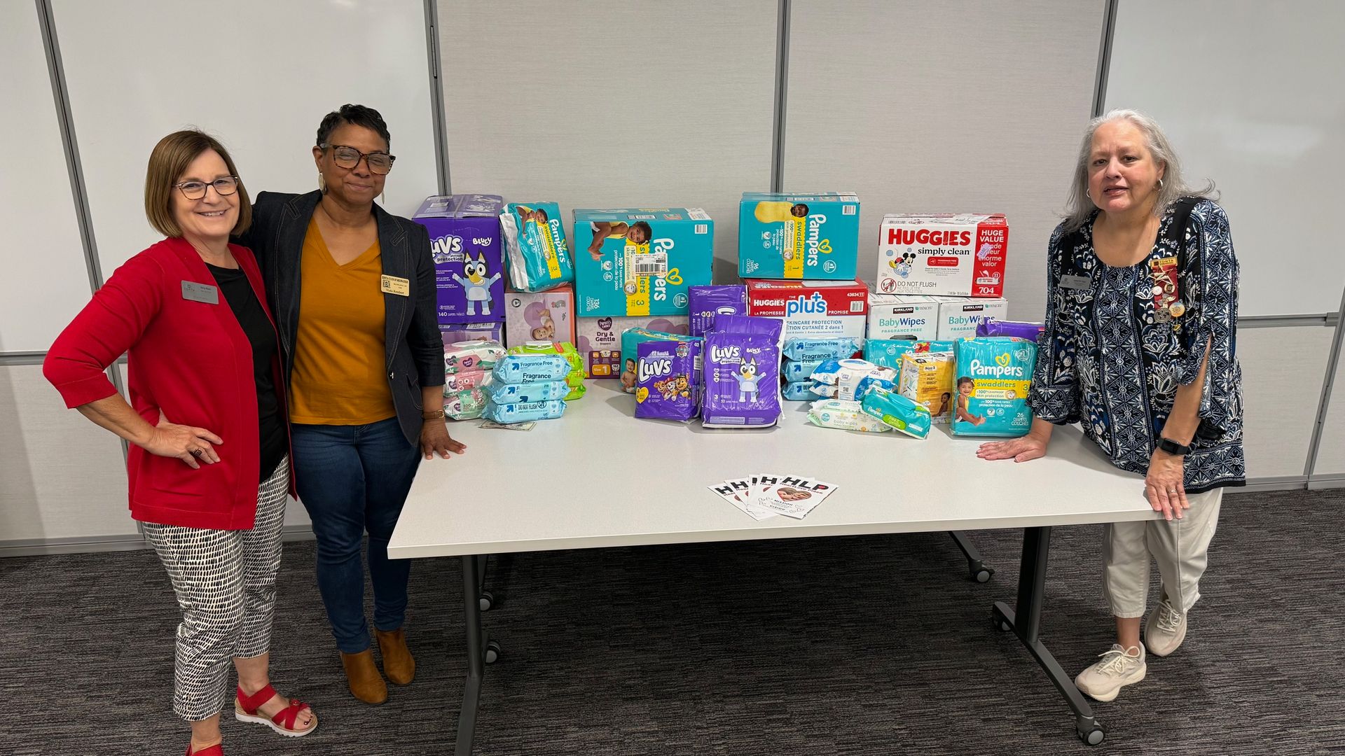 Three women stand by a table of donated diapers and supplies in a room with a whiteboard.