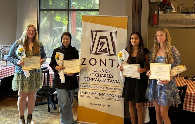 Four women holding certificates and flowers, standing next to a Zonta Club banner indoors.