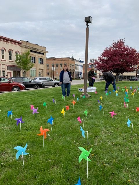 People place colorful pinwheels in a grassy area near a small-town square.