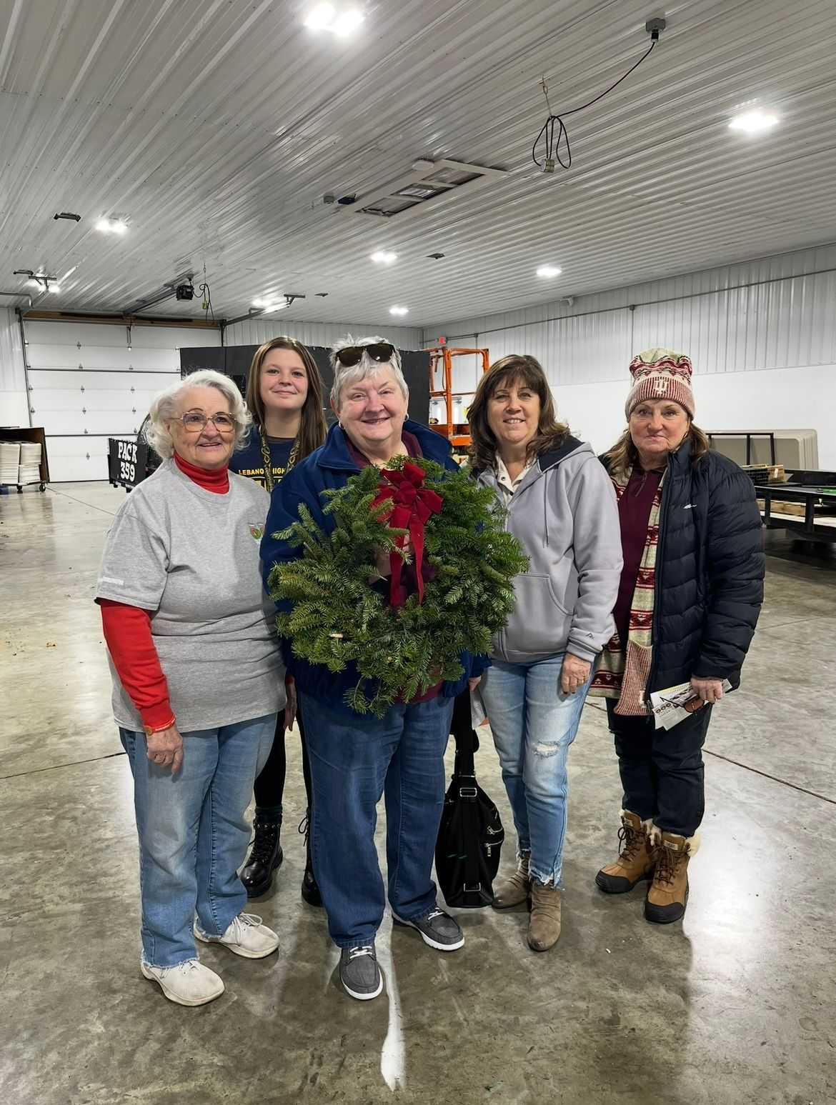 Five people in a large room, one holding a Christmas wreath. They are smiling.