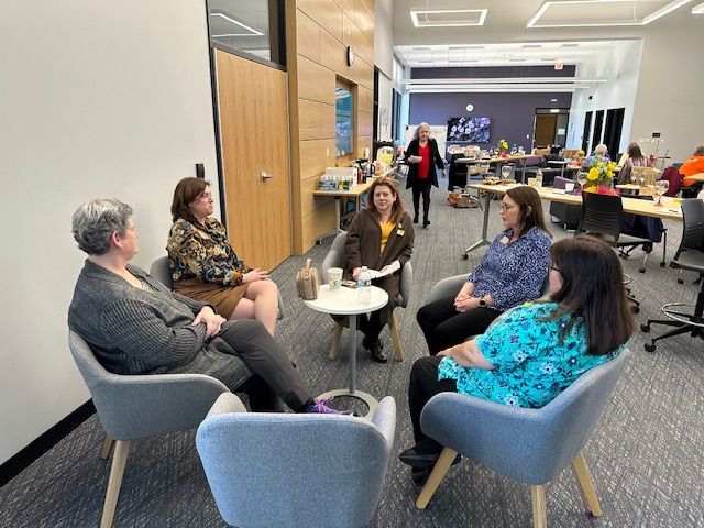 Five people seated in a circle, conversing. An office setting with others working in the background.
