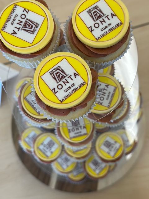 Cupcakes with Zonta Club logo on a glass display. Yellow, brown, and white are the prominent colors.