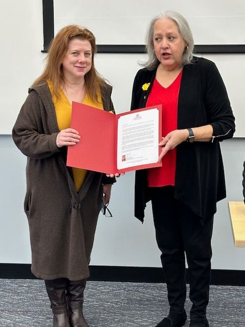 Two women holding a document, one wearing a yellow shirt and brown coat, the other in a red top and black jacket.