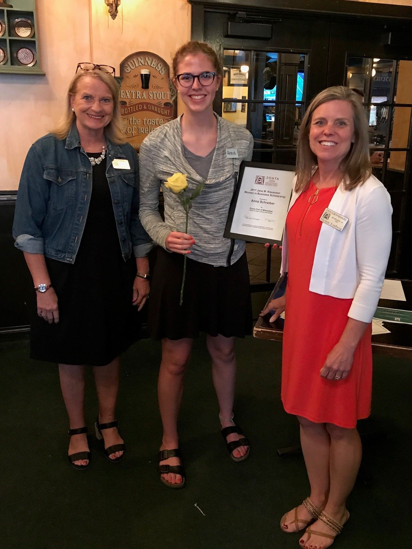 Three women: one holding a framed award, one with a yellow rose, another in a denim jacket, indoors.