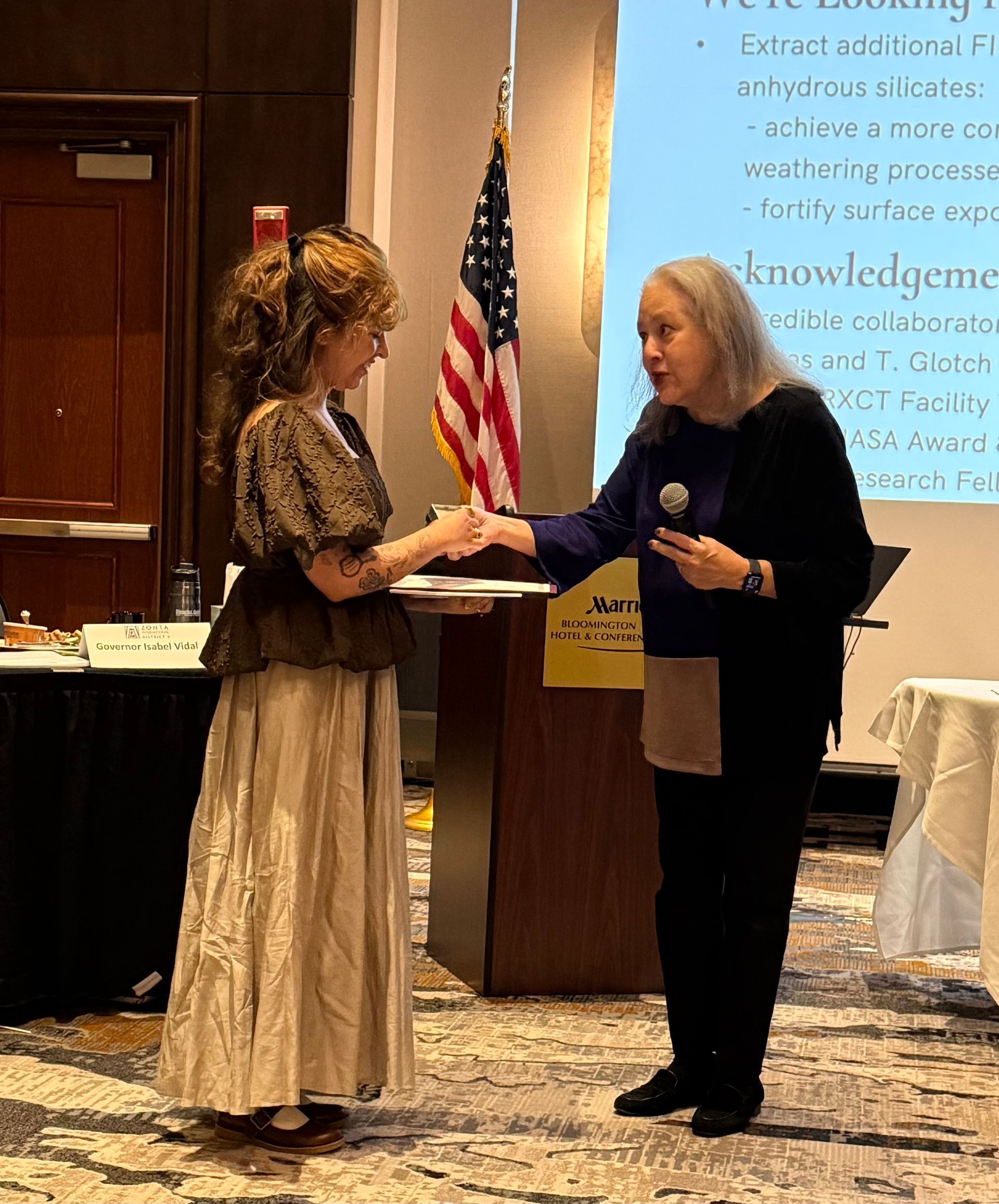 Woman presents a certificate to another woman at an event, U.S. flag visible.