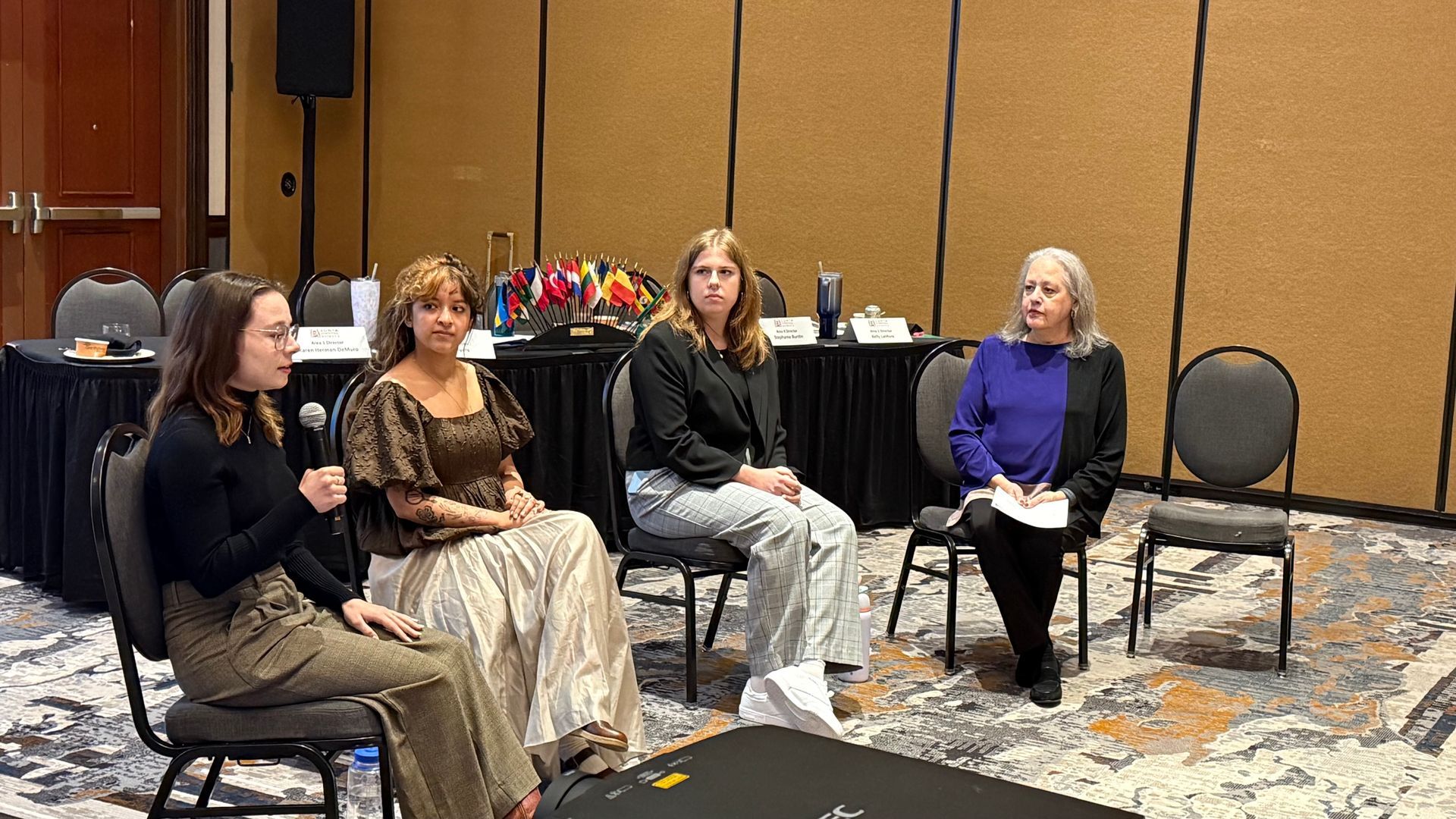Four people on chairs at a table, discussing. Conference room. Microphones and table setting present.