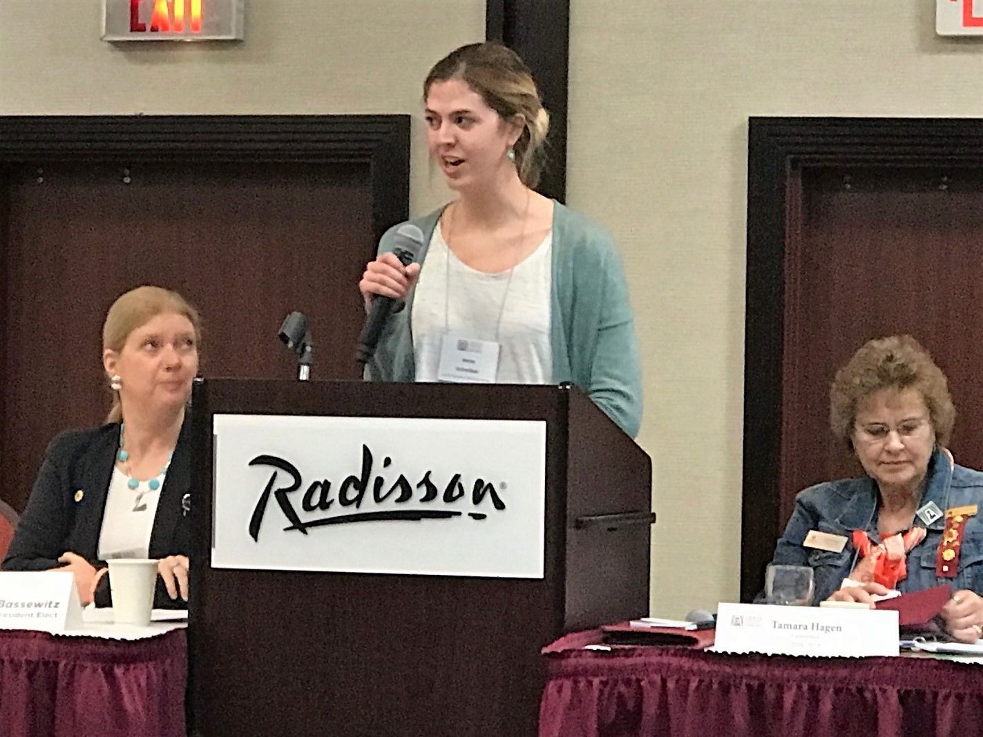 A woman speaks at a Radisson conference podium, flanked by two seated women.