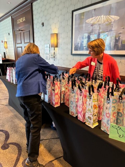 People selecting gift bags from a table, likely at an event. Woman in red jacket assists another. Geneva door visible.