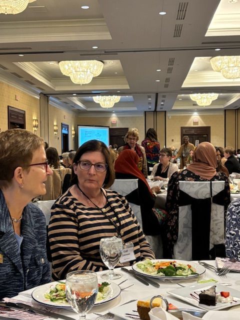 People at tables in a large conference room, eating. Overhead chandeliers and a projector screen are visible.
