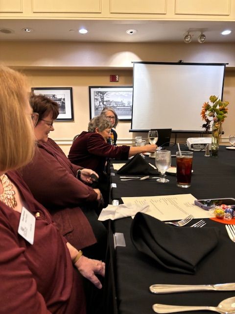 People seated at a table with a projector screen in a banquet hall. Some are holding glasses.