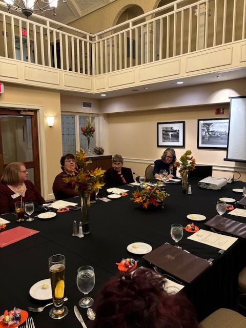 People seated around a black table in a room with a balcony, flowers, and a projector screen.