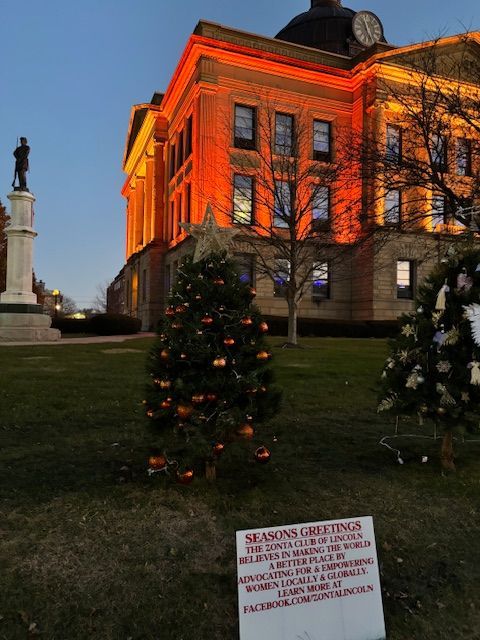 Courthouse with orange lights, Christmas trees, and a sign on a lawn. Statue on the left.