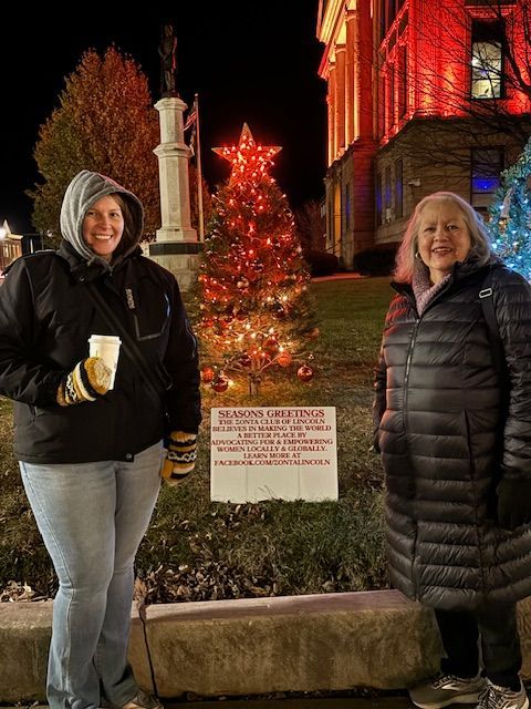 Two women pose near a Christmas tree with a star topper, illuminated building in the background. It's nighttime.
