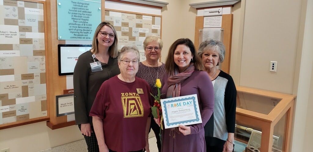 Five women standing in a building, smiling. One woman holds a sign, another a yellow rose.