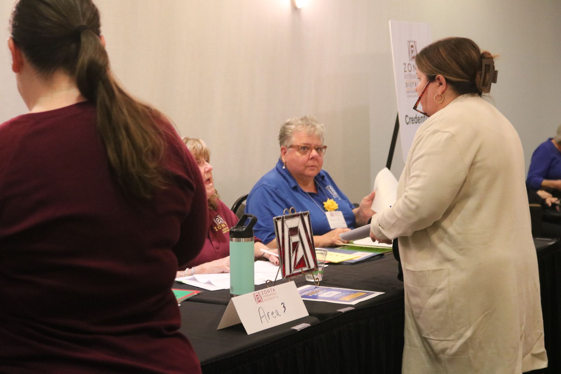 People at a table. Woman in glasses and light coat speaks to others seated behind the table.