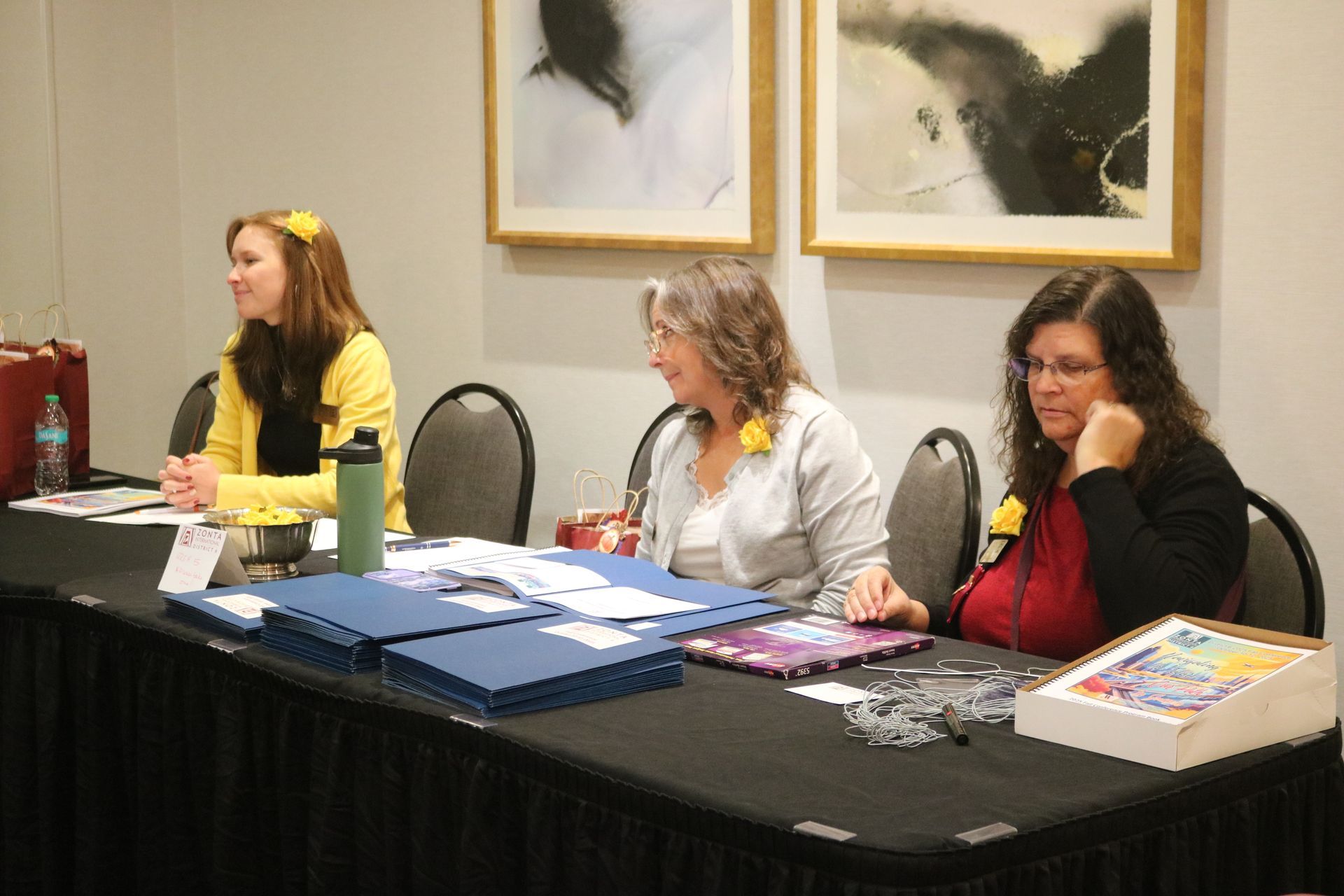 Three people at a table; one wears yellow, one gray, one red. The setting appears to be a conference or event.