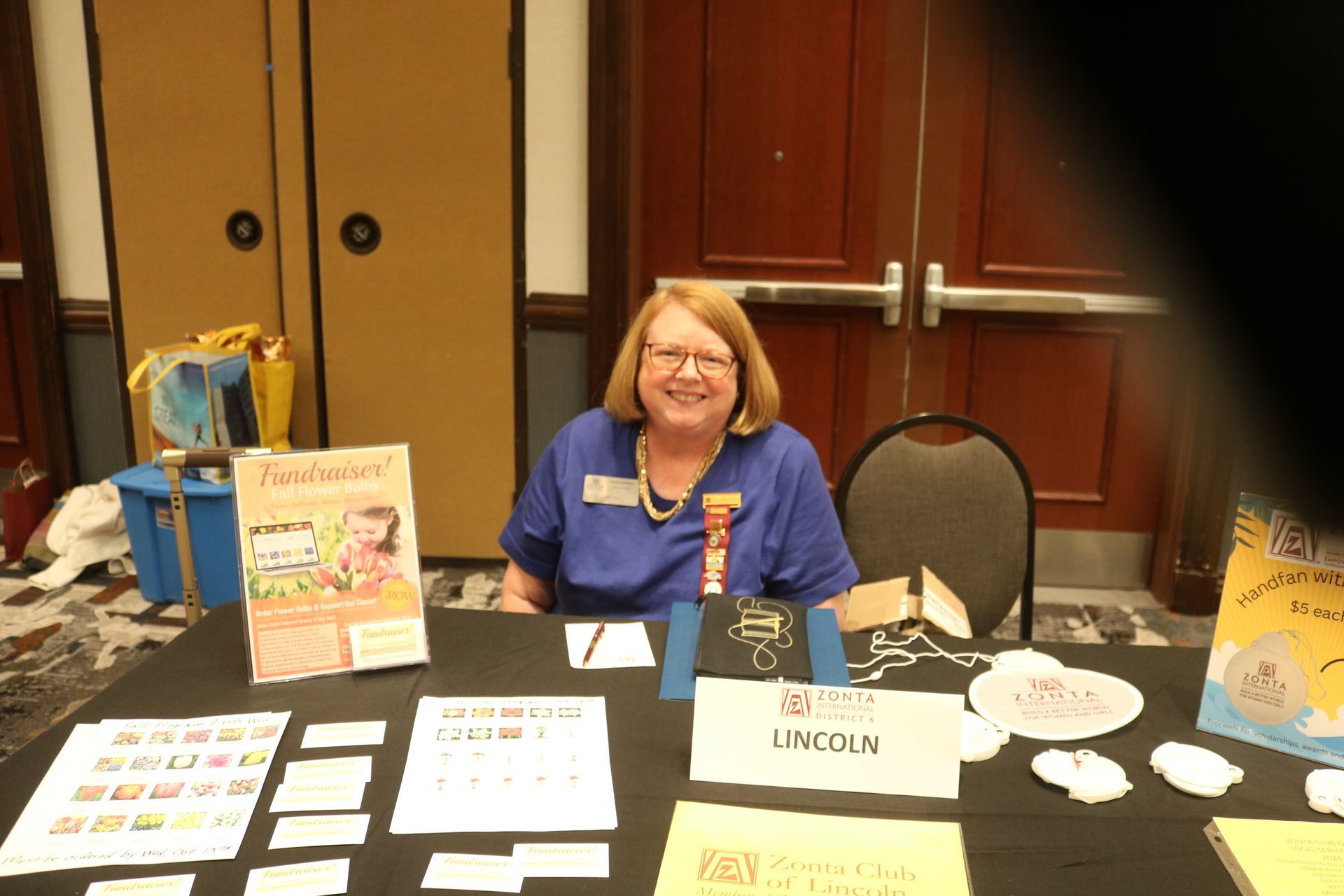 Woman at a table with books and signs, smiling. Interior setting.