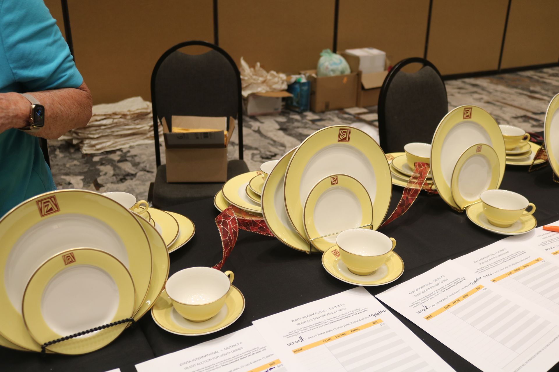Tabletop display of yellow and white china plates, cups, and saucers, with black chairs in the background.