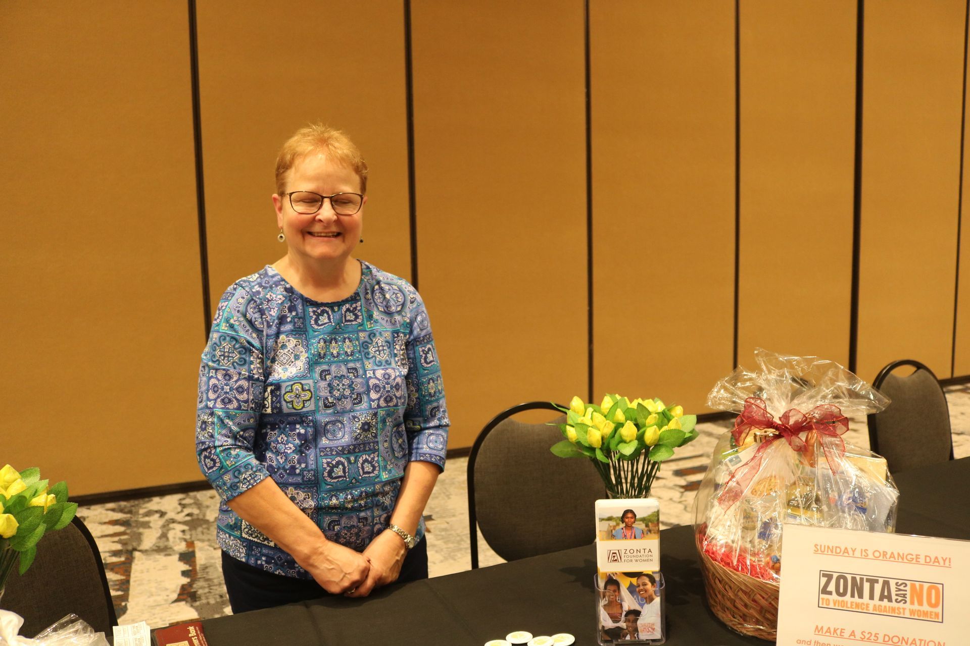 Woman smiling at a booth. There is a gift basket, flowers, and promotional materials on the table. Neutral background.