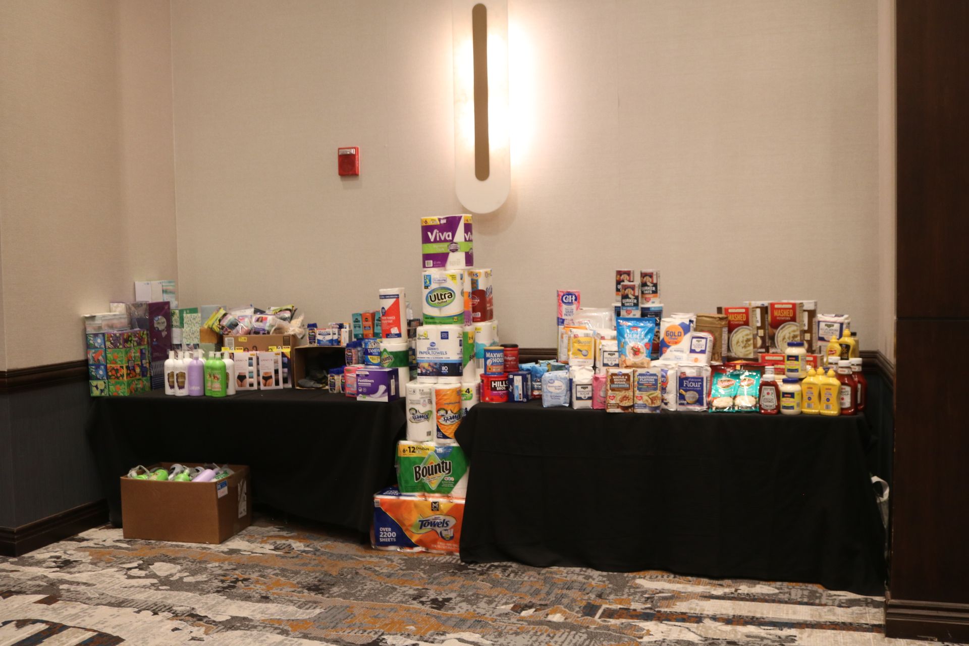 Tables with various packaged food and household items against a wall in a room.