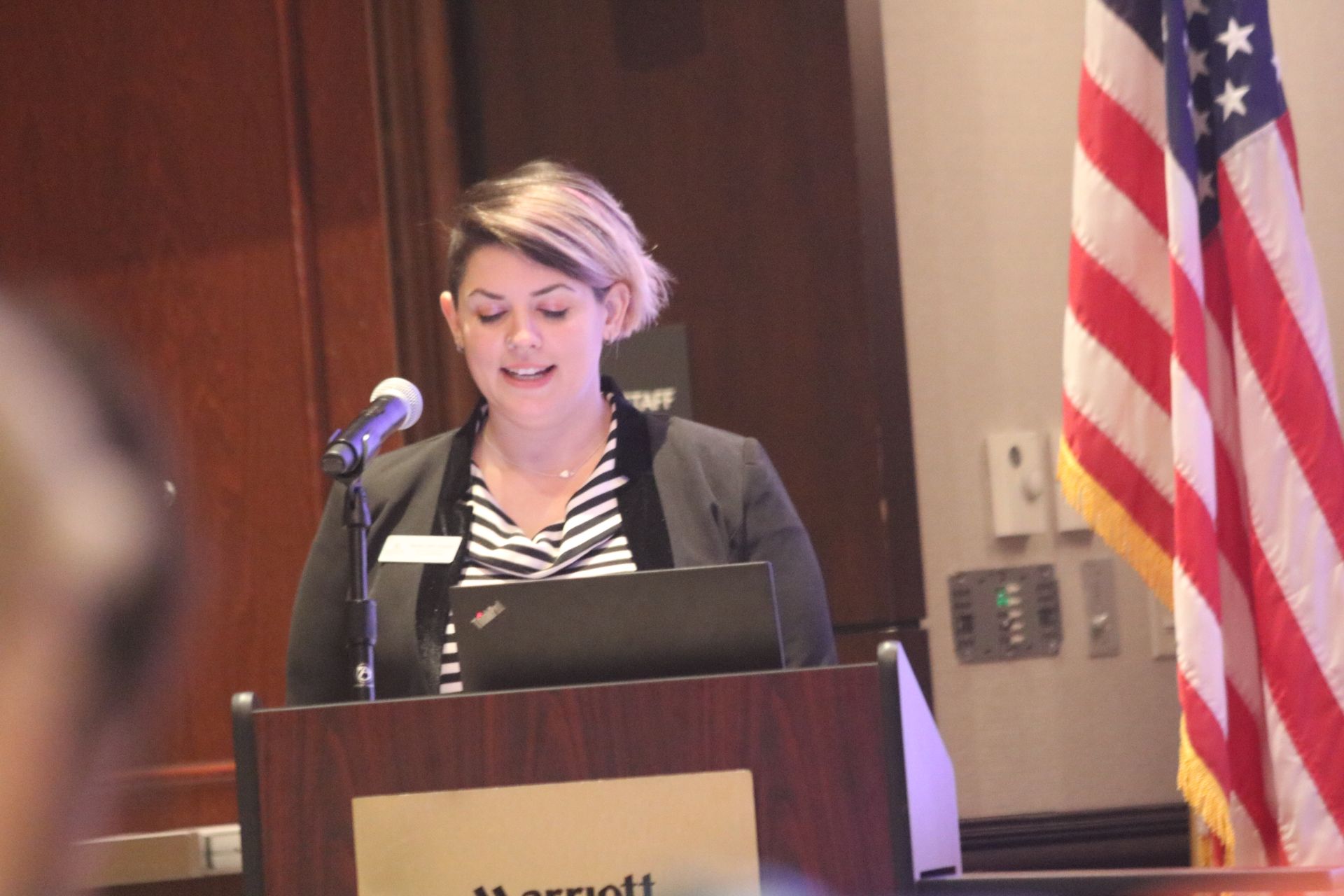 Woman speaking at a podium in front of a US flag, laptop visible. Marriott logo on podium.