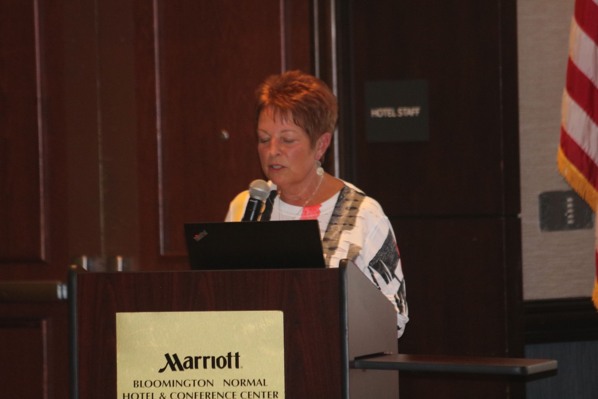 Woman speaking at a Marriott lectern, using a laptop, during a presentation.