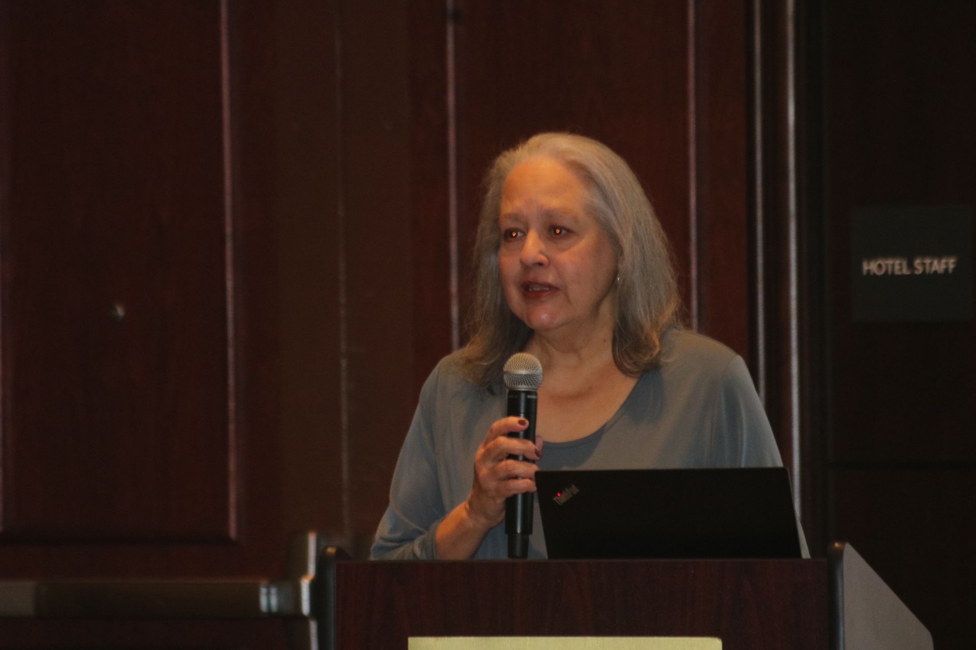 Woman speaking at a podium, holding a microphone. Laptop rests on the podium in a wood-paneled room.