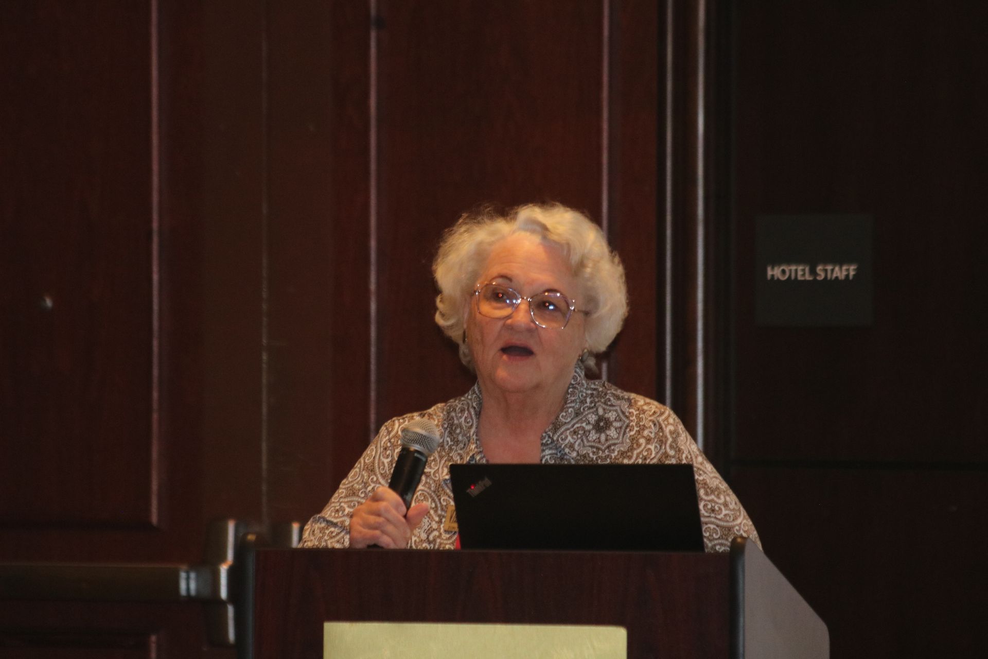 Woman at podium speaking into a microphone. Dark wood background with laptop, 