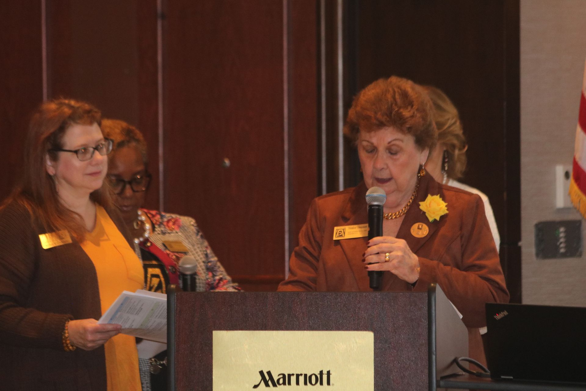 Woman speaks at a Marriott podium, two others stand behind her holding papers.