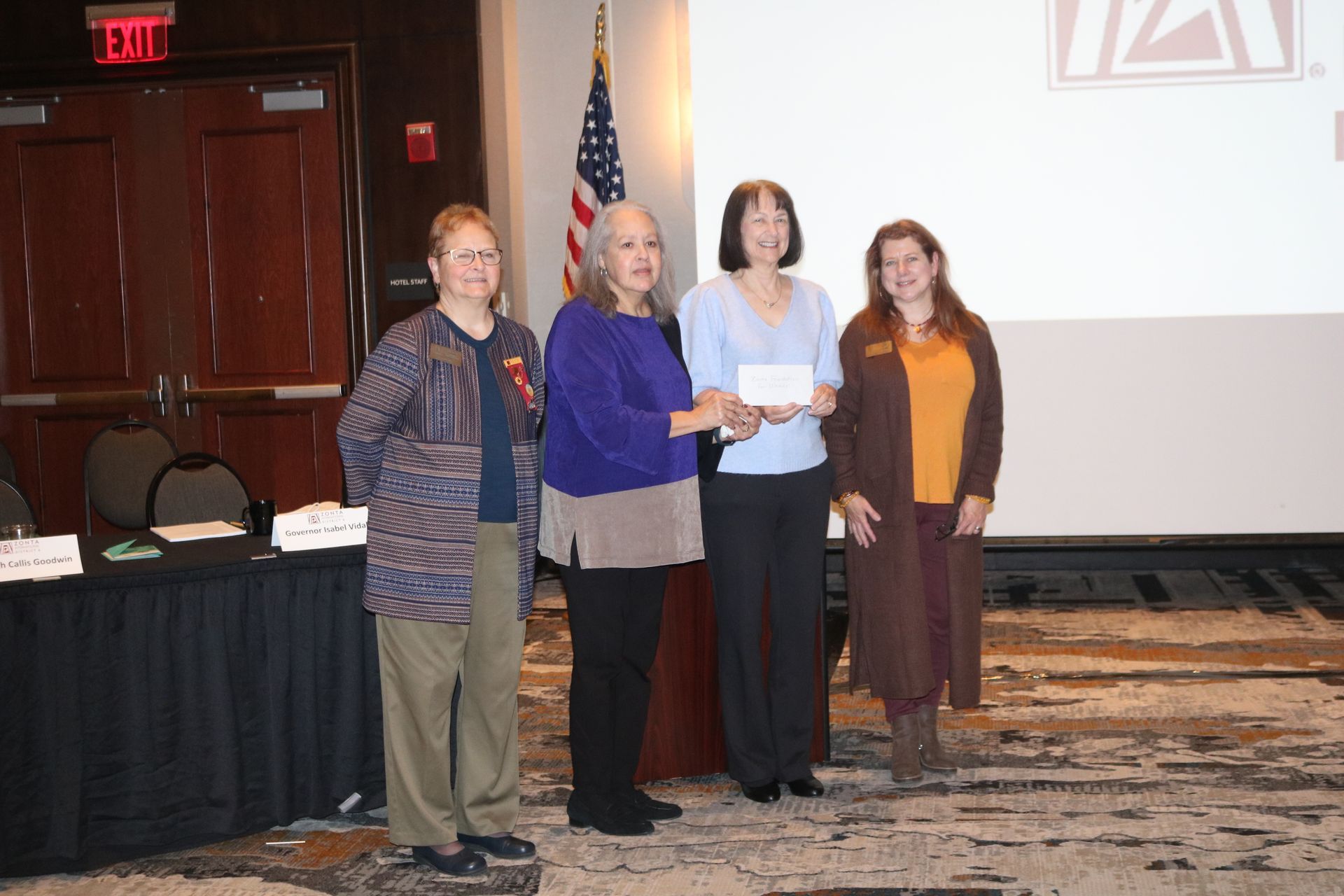 Four people stand on a stage, with one handing a check to another. American flag and a presentation screen in the background.