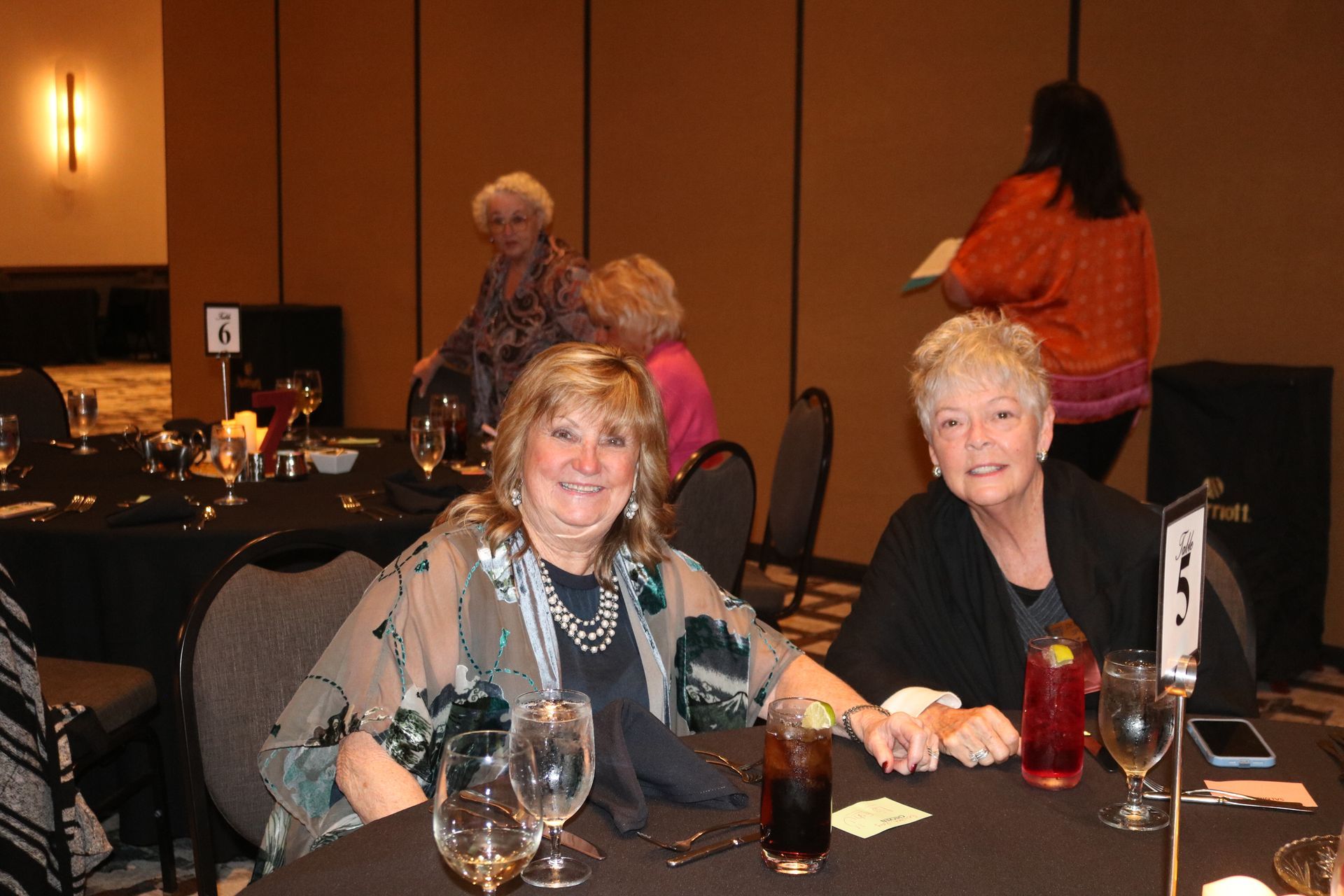 Two women smiling at a table, one in floral top, the other in black, at an event.