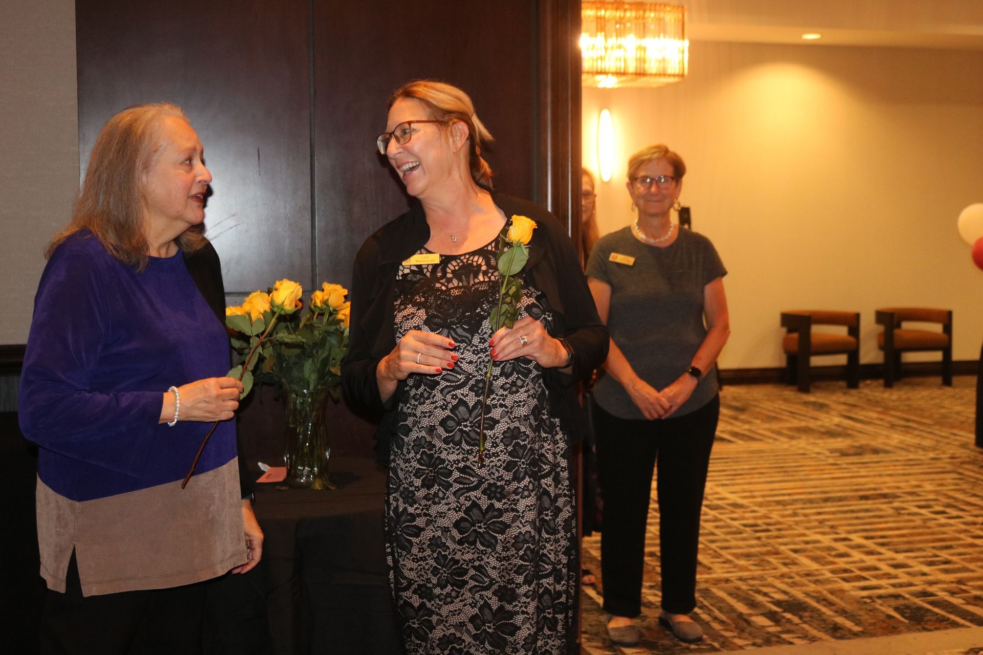 Two women holding yellow roses, smiling, talking near a dark table. Third woman stands in doorway.