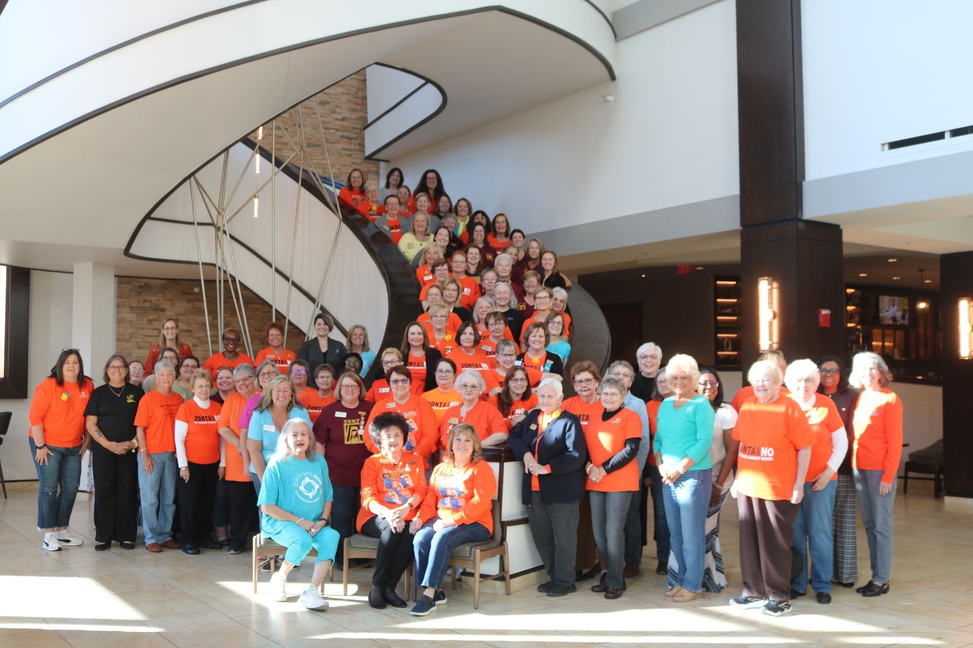 Group of people in orange shirts posing near a modern, curved staircase in a bright lobby.