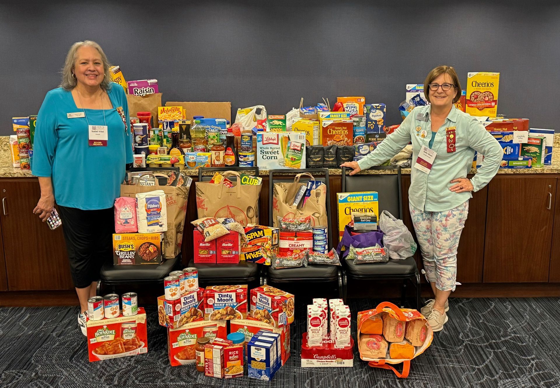 Two women stand with a large food donation; various packaged foods on display, neutral backdrop.
