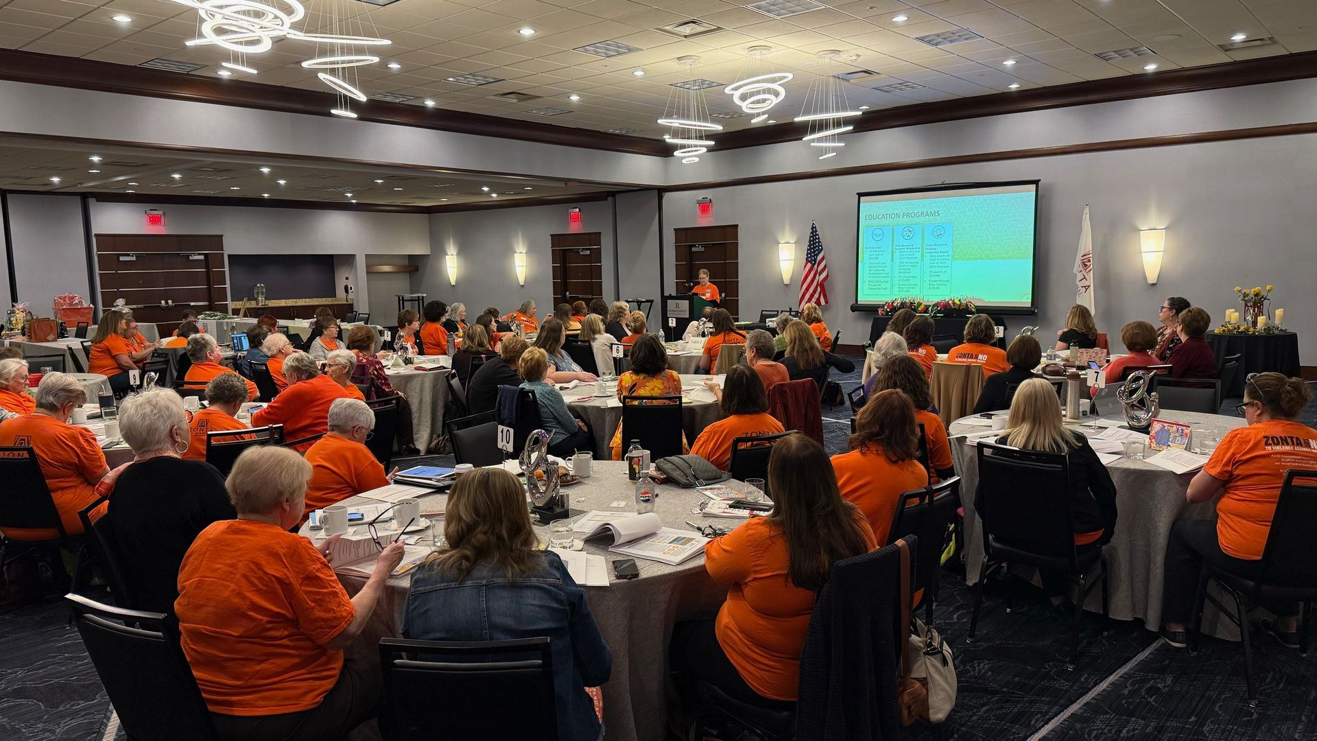 People in orange shirts at a round table event in a banquet hall. A presenter speaks, screen displays data.