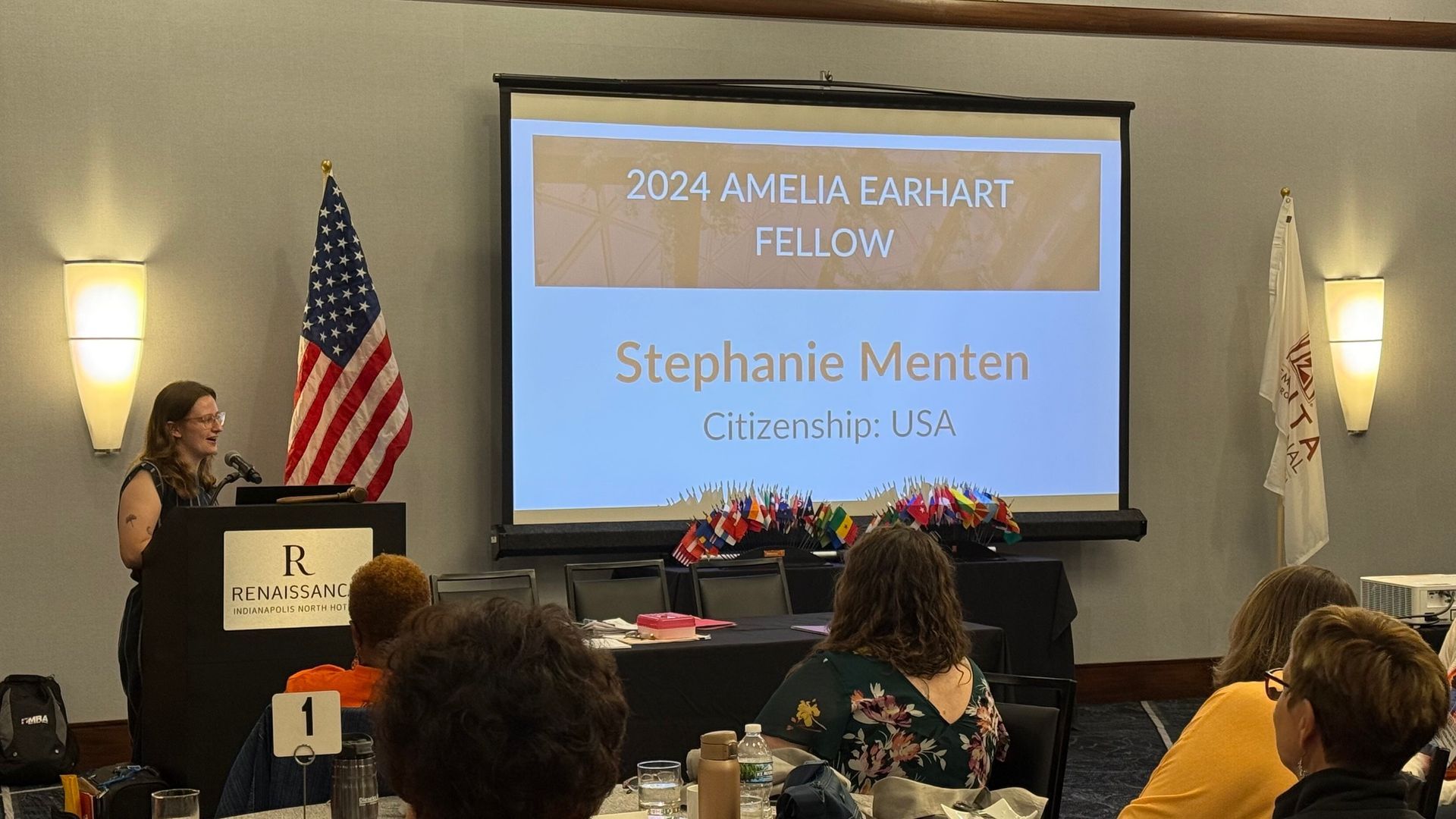 Woman at podium with US flag, presenting at conference. Screen shows