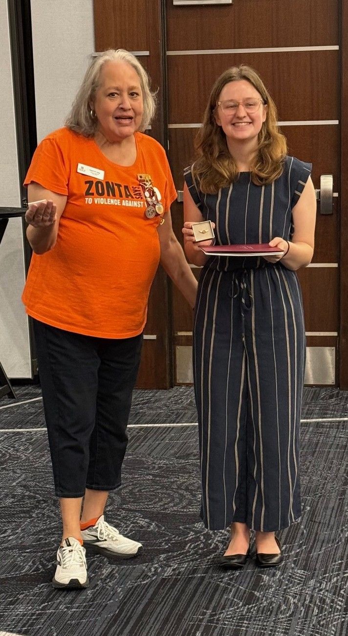 Woman in orange shirt presents award to woman in jumpsuit. Indoors.