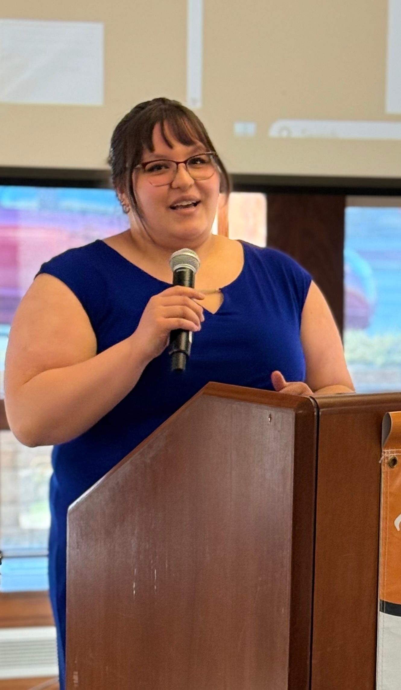 Woman in blue dress speaking at a podium, holding a microphone.