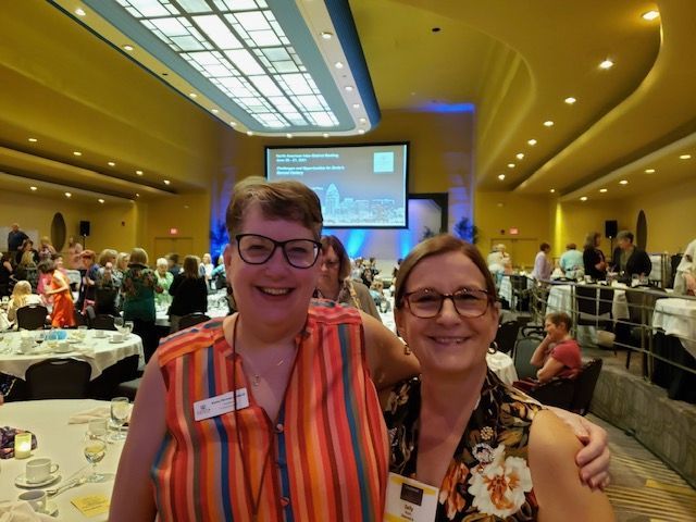 Two women smiling at a conference. Large room with round tables and a screen in the background.