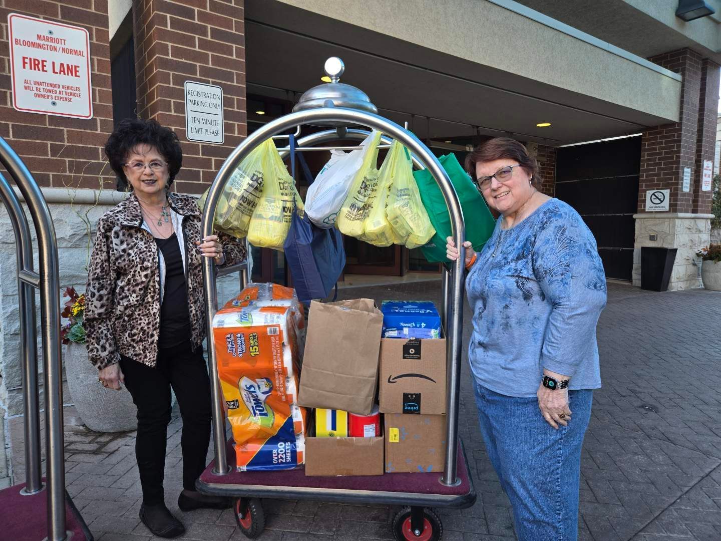 Two women next to a hotel luggage cart loaded with donated supplies, outside a building.
