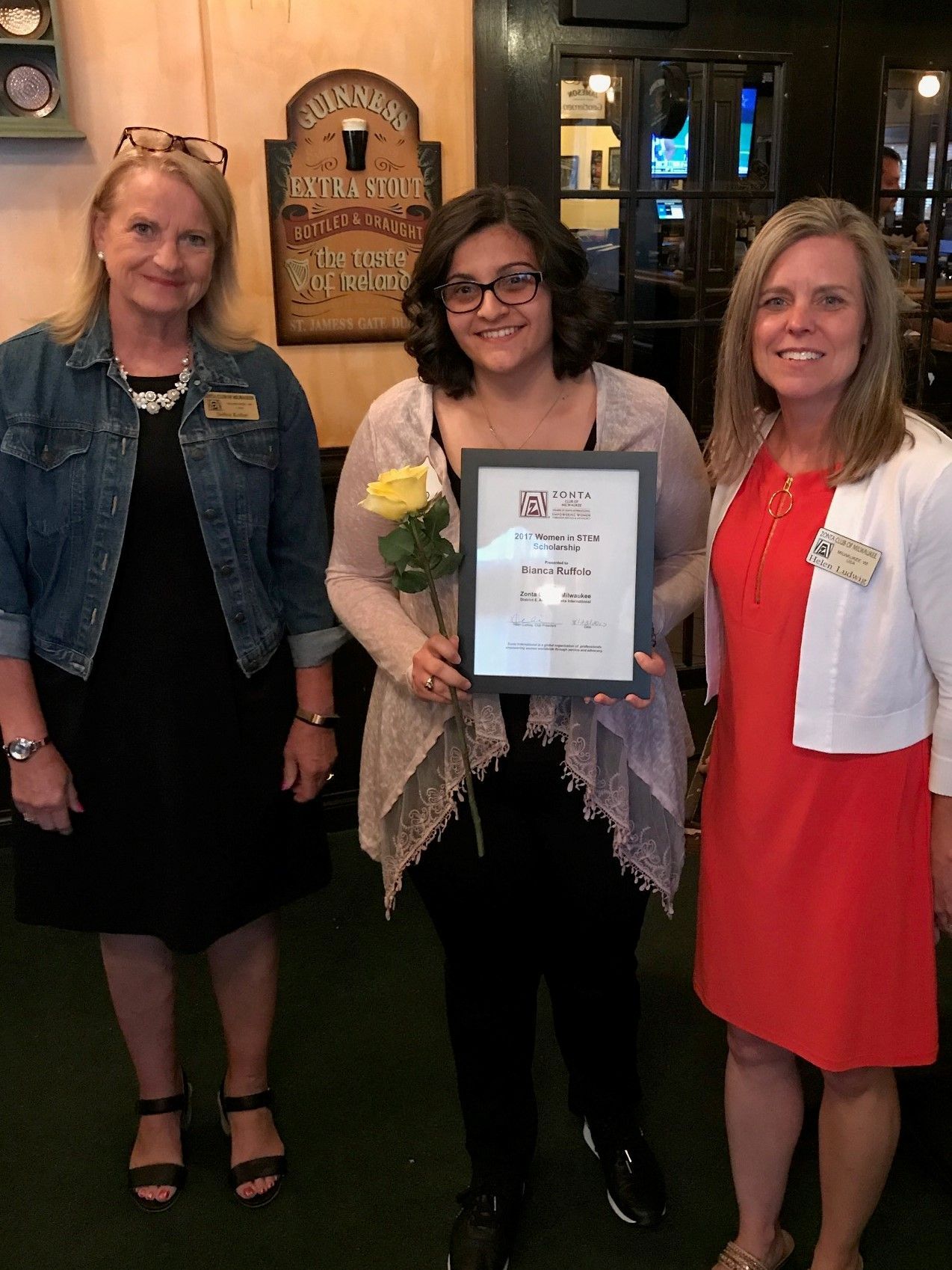 Three women inside a restaurant; a woman in the center holds an award and a yellow rose.