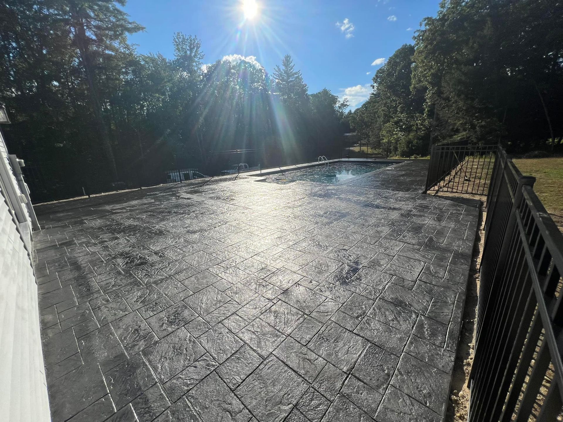 A patio with a pool in the background and the sun shining through the trees