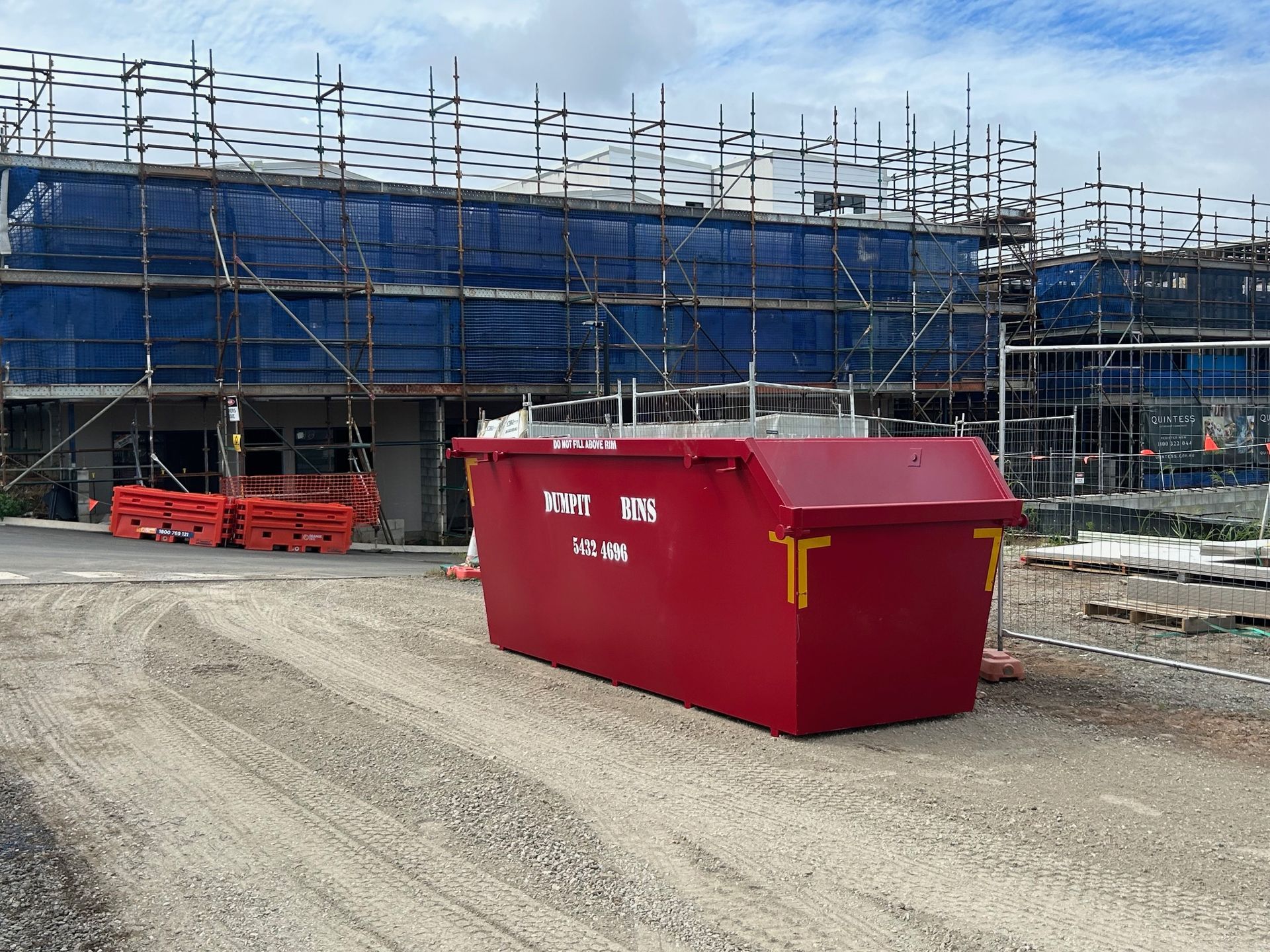 A red Dumpit Handy Bins skip bin sits on a construction site,surrounded by gravel and fencing
