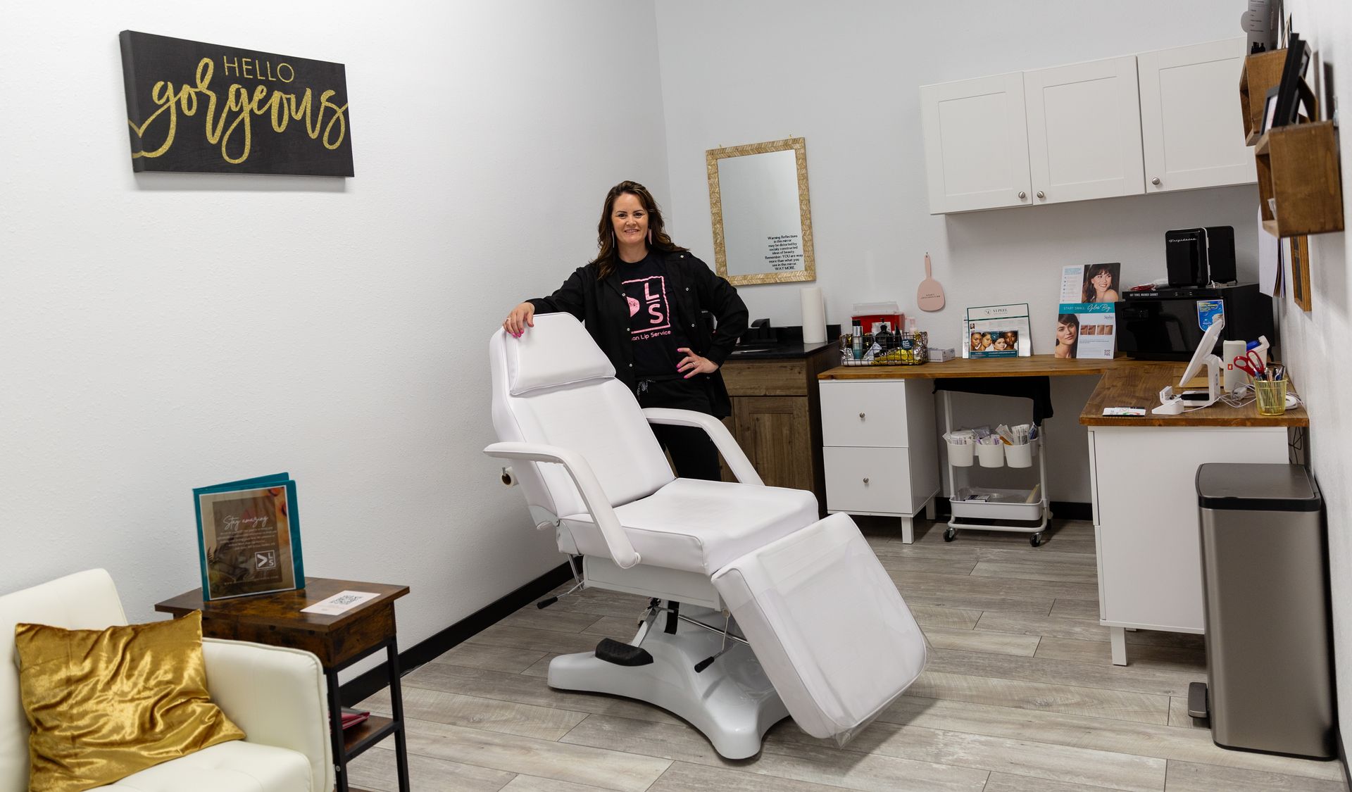 Woman stands near a white treatment chair in a bright beauty treatment room.
