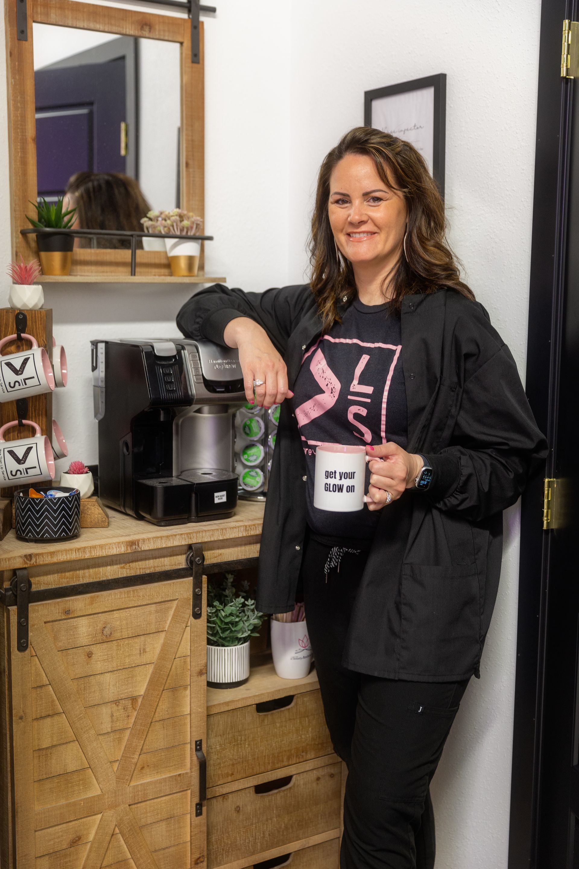 Woman in black jacket holds mug, stands near a coffee machine and wooden cabinet.