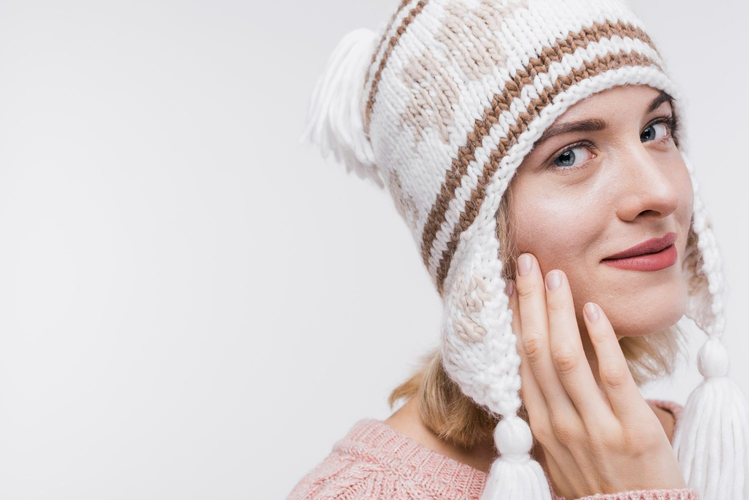 Woman wearing a white and brown knit hat, touching her cheek. Against a white background.