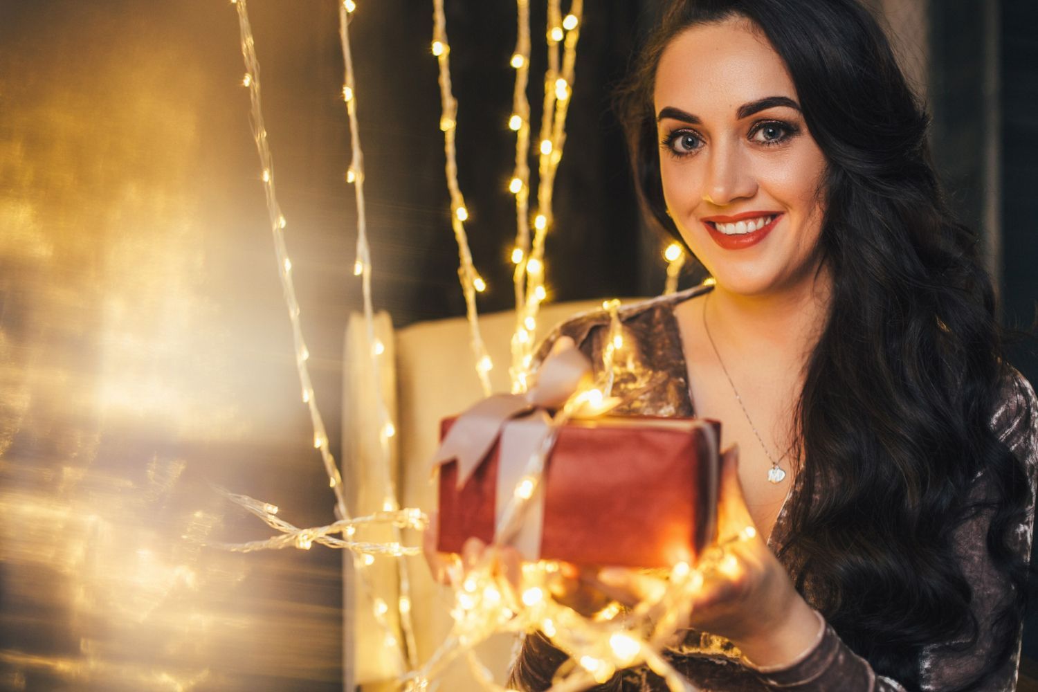 A woman is holding a gift box in her hands surrounded by christmas lights.