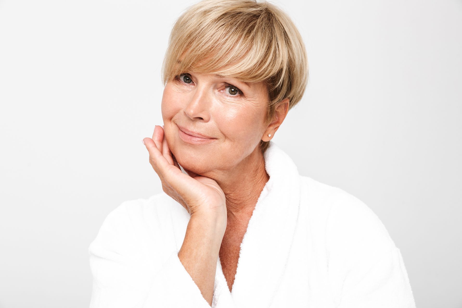 A woman in a white robe is smiling with her hand on her face.