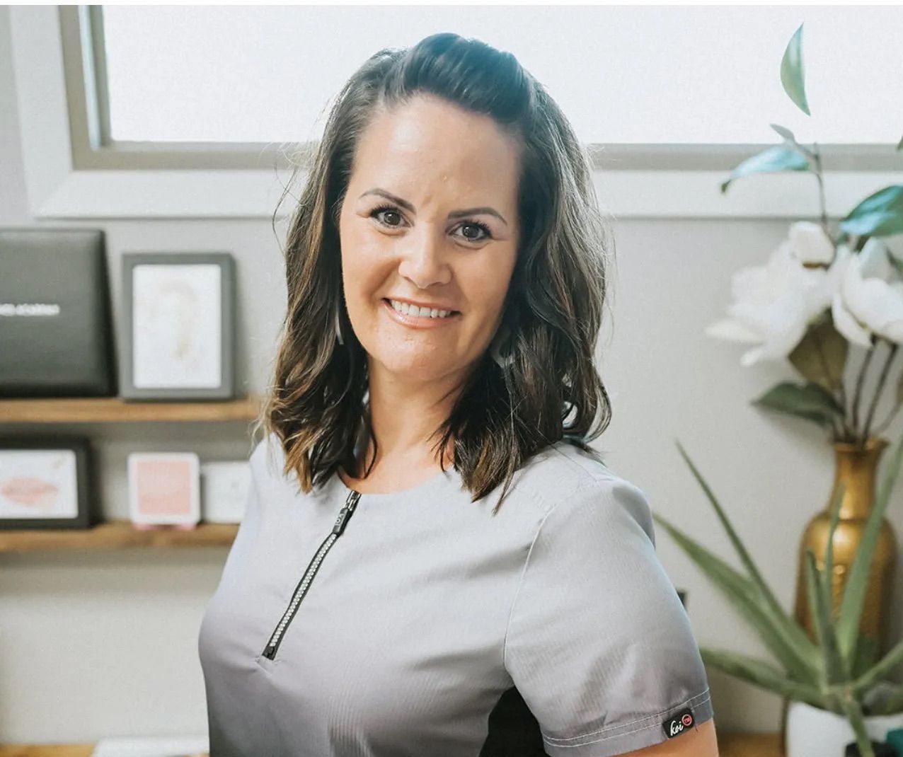 A woman in a grey scrub top is smiling in front of a window.